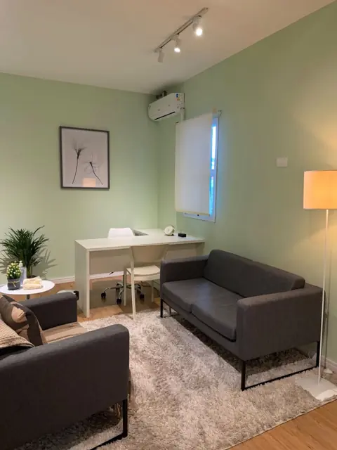 Sala de atendimento com paredes verdes, sofá e poltrona cinza, tapete claro, mesa de trabalho e luminária