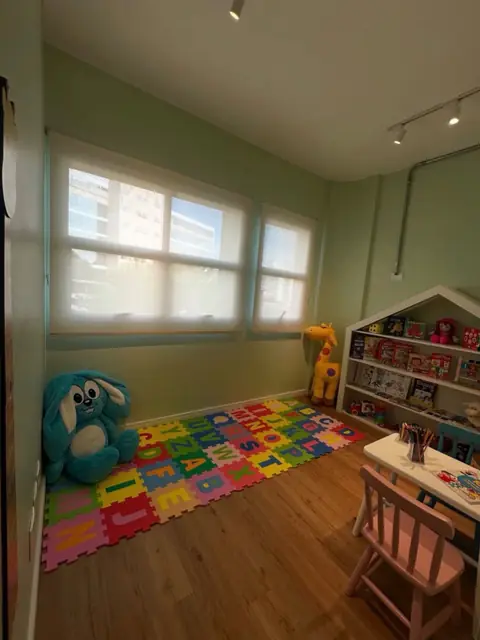Sala infantil com tapete colorido de letras, janela ampla, estante em formato de casinha e mesinha com cadeiras