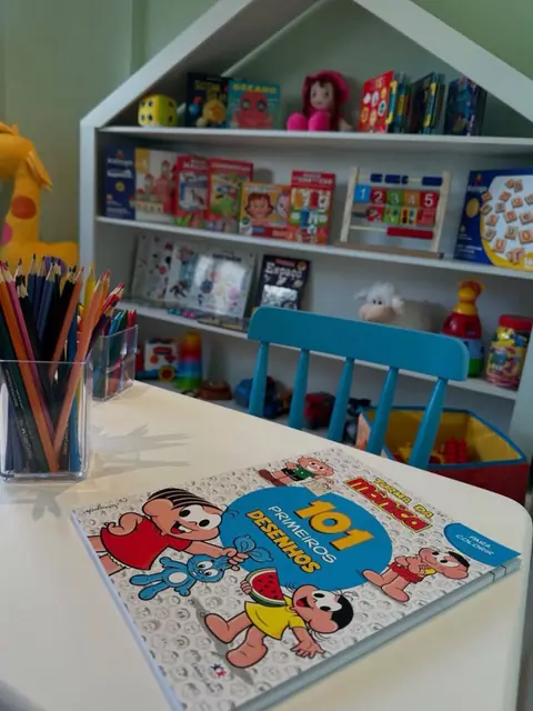 Mesa de atividades com livro de colorir e potes de lápis de cor, com a estante de jogos ao fundo